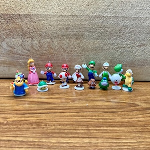May include: A collection of 12 small plastic figurines of characters from the Super Mario Bros. video game franchise. The characters include Mario, Luigi, Peach, Toad, Yoshi, Bowser, and others. The figurines are all in various poses and are painted in bright colors.