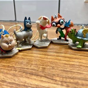 May include: A set of six small plastic figurines of animal characters wearing superhero costumes. The characters include a pig, a dog, a cat, a fox, a turtle, and a bird. Each figurine is on a small base.