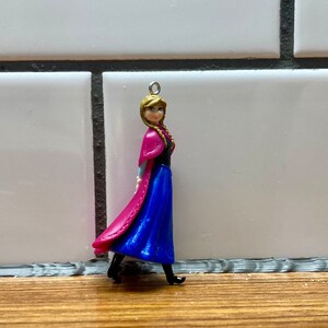 Puede incluir: Una peque&ntilde;a figura de pl&aacute;stico de una ni&ntilde;a con el pelo largo y rubio que lleva un vestido azul y una capa rosa. Lleva zapatos negros y tiene un lazo plateado en la parte superior de la cabeza.