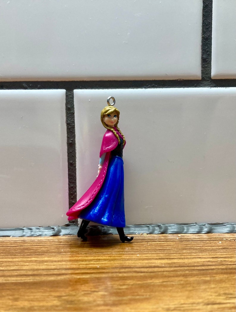 Puede incluir: Una peque&ntilde;a figura de pl&aacute;stico de una ni&ntilde;a con el pelo largo y rubio que lleva un vestido azul y una capa rosa. Lleva zapatos negros y tiene un lazo plateado en la parte superior de la cabeza.