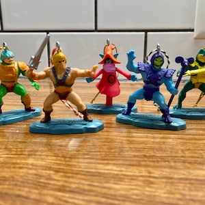 May include: Set of six He-Man and the Masters of the Universe Christmas ornaments. The ornaments are made of plastic and feature characters from the popular cartoon series. The characters include He-Man, Skeletor, Beast Man, Man-At-Arms, Teela, and Evil-Lyn. Each ornament is suspended from a gold string.