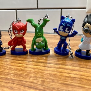 May include: Five small plastic figurines of characters from the children's television show 'PJ Masks'. The characters are Gekko, Owlette, Catboy, Romeo, and Night Ninja. Each figurine is attached to a string and has a blue base.