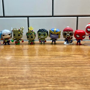 Puede incluir: Una colección de nueve figuras de superhéroes de Marvel con temática navideña. Las figuras están todas vestidas con atuendos festivos, incluidos gorros de Papá Noel, cuernos de reno y luces navideñas. Las figuras están dispuestas en fila sobre una superficie de madera.