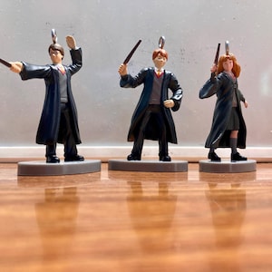 Może przedstawiać: Trzy miniaturowe figurki postaci z serii fantasy. Każda figurka stoi na szarej podstawie i trzyma różdżkę. Mają na sobie czarne szaty i srebrną pętlę u góry do zawieszenia. Figurki to Harry Potter, Ron Weasley i Hermiona Granger.