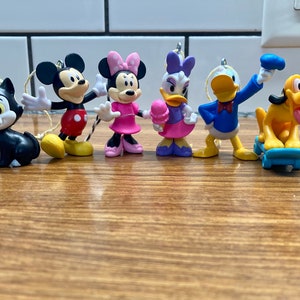May include: A set of seven small plastic Disney character ornaments. The characters include Mickey Mouse, Minnie Mouse, Donald Duck, Goofy, Pluto, and two others. The ornaments are all in different colors and have a variety of expressions.
