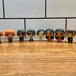 May include: A collection of 11 Harry Potter character figurines, including Harry Potter, Ron Weasley, Hermione Granger, and others. The figurines are all in the popular Funko Pop! style, with large heads and small bodies. They are all standing on a wooden surface.
