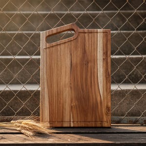 Puede incluir: Una tabla de cortar rectangular de madera con asa. La tabla es de madera marrón claro con vetas visibles. Una ranura recorre los bordes. El asa es un recorte en la parte superior. Espigas de trigo en primer plano.