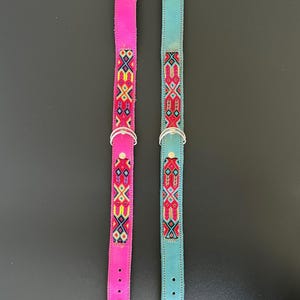 May include: Two dog collars. One is bright pink, the other is teal. Both have a silver metal buckle and a D-ring. The collars are decorated with a colorful embroidered pattern in red, yellow, and blue. The collars are made of leather.