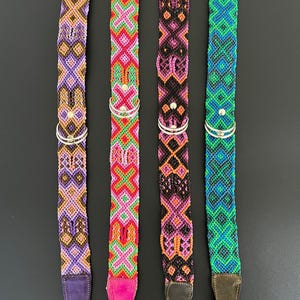 May include: Four colorful dog collars with intricate woven patterns. Each collar features a different color scheme: purple, pink, black, and blue. They have silver metal hardware and leather ends. The collars are designed for pets.