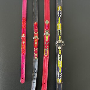 May include: Four leather dog collars in varying colors. The collars are pink, black, red, and black. Each collar has a silver buckle and a decorative beaded design in various colors. The collars are displayed on a black surface.