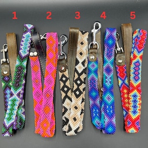 May include: A collection of six colorful dog leash handles with woven patterns. Each handle features a different design, including geometric and diamond patterns, in various colors like green, pink, blue, and red. They have brown leather accents and metal clasps.