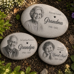 May include: Three oval-shaped light gray memorial stones featuring engraved black portraits and text. One stone reads Grandma 1948-2022, another reads Grandpa 1944-2021, and the third reads Sweet Angel 2019-2020 with various memorial phrases.
