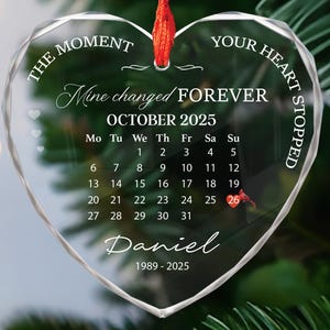 May include: Clear heart-shaped ornament with the text "The Moment Your Heart Stopped" and "Mine changed FOREVER". It features a calendar for October 2025 and the name "Daniel" with the years 1989-2025. A red ribbon is attached for hanging.