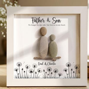 Könnte beinhalten: Ein gerahmtes Kunstwerk mit einem Vater und Sohn aus Steinen. Der weiße Rahmen umgibt einen weißen Hintergrund mit dem Text "Father & Son" und "No longer by my side, but forever in my heart." Pusteblumen sind unten.