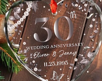 May include: A heart-shaped glass ornament with the text "30th Wedding Anniversary Oliver & Jenny 11.25.1995". The ornament has a red ribbon for hanging and is decorated with a butterfly and floral accents. The background is a wooden surface with Christmas decorations.