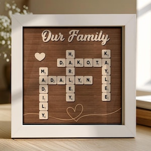 May include: A framed wooden family sign with the words "Our Family" in cursive. The sign features wooden letter tiles spelling out names, a heart, and a decorative heart outline. The frame is white, and the background is a dark wood grain.