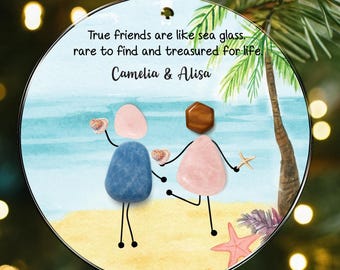 May include: A round ceramic ornament featuring a beach scene with two figures made of smooth stones. The text reads: True friends are like sea glass, rare to find and treasured for life, Camelia & Alisa. It has a red and white gingham bow.