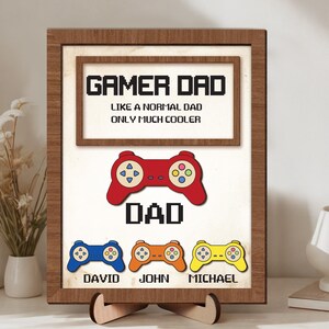 May include: A wooden framed sign with the text "GAMER DAD" and "LIKE A NORMAL DAD ONLY MUCH COOLER". Below is a red controller graphic and the word "DAD". Three controllers are below with the names David, John, and Michael.