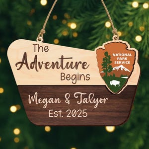 May include: Wooden sign with the text "The Adventure Begins" and "Megan & Talyor Est. 2025". Features a National Park Service emblem. The sign is two-toned, with a light wood top and a dark wood bottom, and is suspended by a twine string.