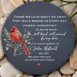 Könnte beinhalten: Ein runder, dunkelgrauer Gedenkstein aus Schiefer mit einer Illustration eines roten Kardinals und Text. Der Text lautet: "Die, die wir lieben, gehen nicht weg... In liebevoller Erinnerung an John Redfield 1958-2024".