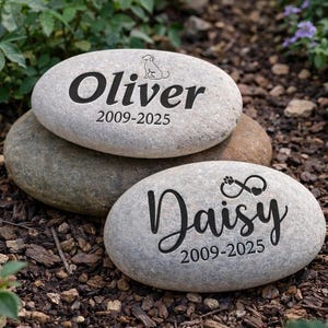 May include: Three gray engraved stones with the names "Oliver" and "Daisy" in black script. The "Oliver" stone has a dog silhouette, and the "Daisy" stone has a paw print with an infinity symbol. Each stone includes dates.