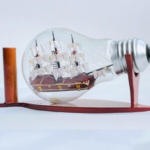 May include: A decorative ship in a bottle, displayed on a wooden stand. The ship is brown with white sails, encased in a clear glass lightbulb. The stand is a reddish-brown colour.