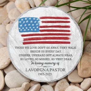 May include: A circular memorial stone featuring an American flag design. The flag is red, white, and blue. Text reads "Those we love don't go away... In loving memory of LAVORGNA PASTOR 1985-2025."