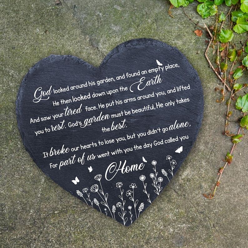 May include: A heart-shaped, black slate memorial stone with white script. The text reads, "God looked around his garden...He only takes the best...Home." Delicate floral and butterfly designs accent the bottom edge. A touching garden decor item.