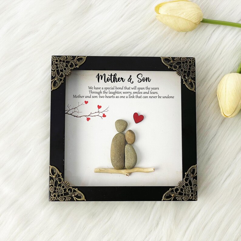 May include: A framed artwork featuring a pebble art design of a mother and son holding hands. The artwork is set against a white background with a black frame and ornate gold corners. The text reads "Mother & Son" and "We have a special bond that will span the years Through the laughter, worry, smiles and tears. Mother and son: two hearts as one a link that can never be undone."
