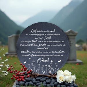 May include: A round, dark gray memorial stone with white script, floral accents, and a butterfly design. The stone is placed on a bed of black stones, with white roses and red berries in the foreground. The background features a green field and mountains.