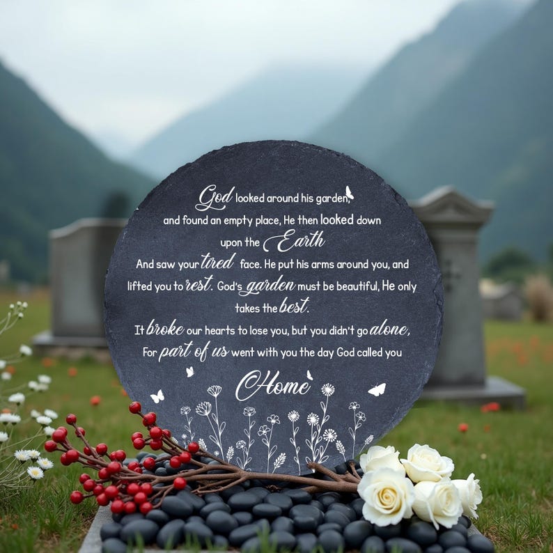 May include: A round, dark gray memorial stone with white script, floral accents, and a butterfly design. The stone is placed on a bed of black stones, with white roses and red berries in the foreground. The background features a green field and mountains.