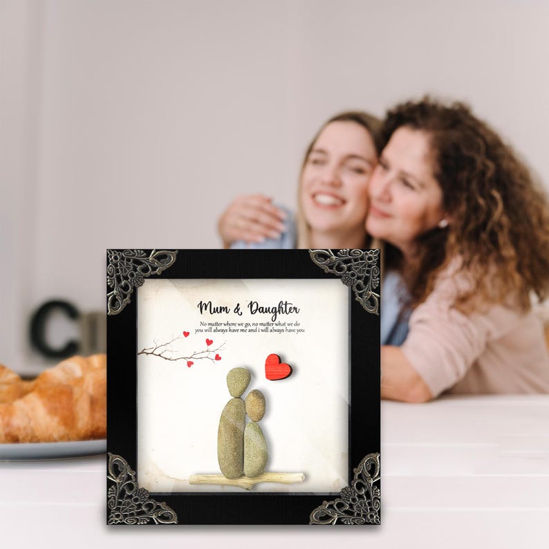 Puede incluir: Una pieza de arte de guijarros enmarcada con un marco negro y esquinas plateadas ornamentadas. El arte representa dos figuras hechas de guijarros, que representan a una madre y una hija, con un coraz&oacute;n rojo sobre ellas. El texto dice "Mam&aacute; e Hija" y "No importa a d&oacute;nde vayamos, no importa lo que hagamos, siempre me tendr&aacute;s y siempre te tendr&eacute;."