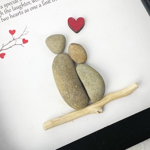 May include: A framed artwork featuring a couple made of stones, sitting on a branch with a red heart above them. The artwork has a white background and a black frame. The text "We have a special... Through the laughter, we... her and son: two hearts as one a link..." is visible on the white background.
