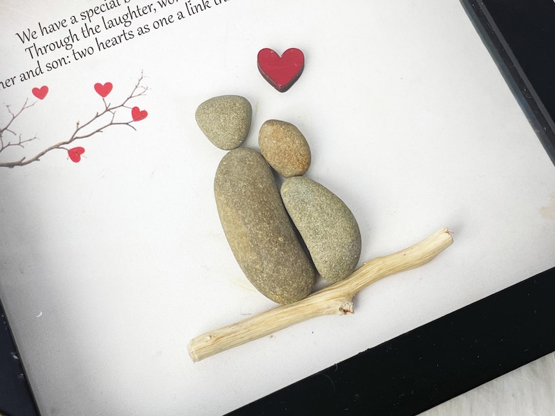 May include: A framed artwork featuring a couple made of stones, sitting on a branch with a red heart above them. The artwork has a white background and a black frame. The text "We have a special... Through the laughter, we... her and son: two hearts as one a link..." is visible on the white background.