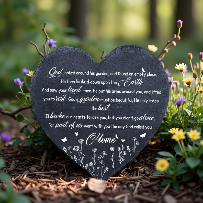 May include: A heart-shaped, dark gray stone memorial plaque with white script. The inscription includes the words "God," "Earth," and "Home," along with a floral and butterfly design. The plaque is surrounded by flowers.