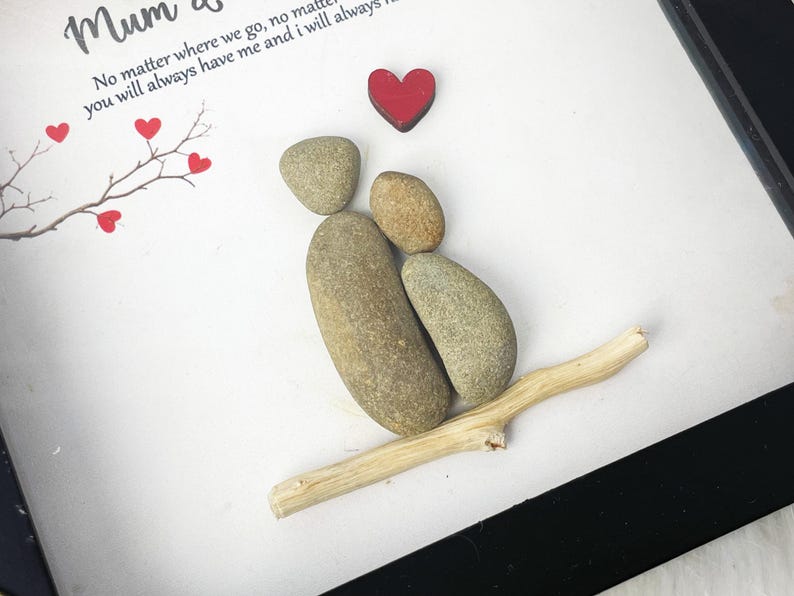 Puede incluir: Una obra de arte enmarcada que presenta una pareja hecha de piedras, sentada en una rama con un coraz&oacute;n rojo sobre ellos. La obra de arte tiene el texto "Mum &" y "No matter where we go, no matter you will always have me and I will always" sobre un fondo blanco.
