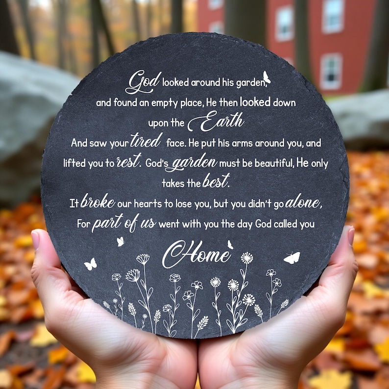 May include: A round, dark gray stone plaque with white script and floral accents. The text includes the words "God," "Earth," and "Home." The plaque is held in two hands against a blurred background of fall foliage.