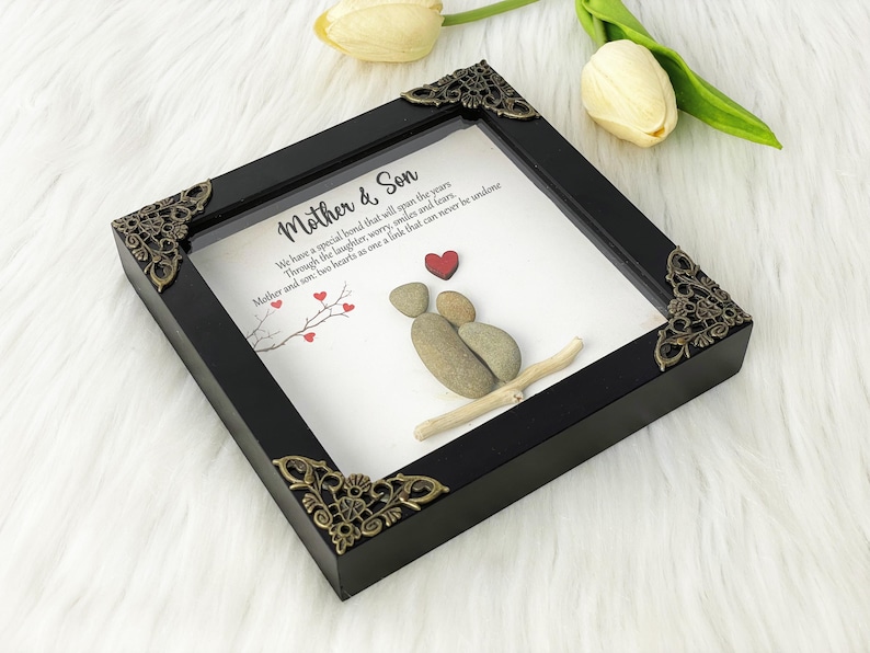 May include: A framed artwork featuring a pebble art design of two figures holding hands, representing a mother and son. The artwork is set against a white background with a quote that reads: "We have a special bond that will span the years. Through the laughter, worry, smiles and tears. Mother and son, two hearts as one, a link that can never be undone."