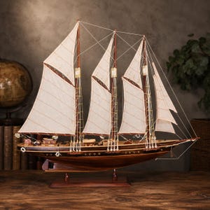 May include: A wooden model ship featuring a dark brown hull, three tall masts, and off-white fabric sails secured with thin rigging lines. The vessel is mounted on a rectangular wooden base and includes small deck details like cabins and life rings.