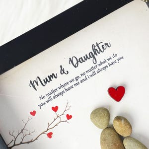 Puede incluir: Una impresi&oacute;n enmarcada con un marco negro y un fondo blanco. La impresi&oacute;n presenta una cita sobre el v&iacute;nculo entre una madre y una hija, junto con un peque&ntilde;o &aacute;rbol con corazones rojos y un grupo de piedras grises. La cita dice: "Mam&aacute; e Hija No importa a d&oacute;nde vayamos, no importa lo que hagamos, siempre me tendr&aacute;s y yo siempre te tendr&eacute;."