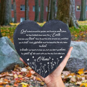 May include: A heart-shaped, dark gray slate memorial stone with white script and floral accents. The text includes a poem about loss and the word "Home". The stone is held in a hand against a blurred outdoor background.