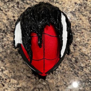 May include: A red and black Spider-Man mask with black dripping paint on the top and white accents on the sides.