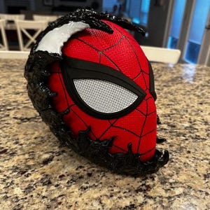 May include: A red and black Spider-Man mask with a black Venom symbiote design. The mask has a black mesh eye piece and a white spider logo.