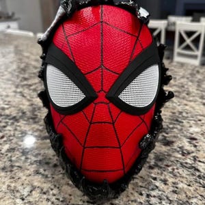 May include: A red and black Spider-Man mask with a black and white mesh eye design. The mask is surrounded by a black, textured, jagged edge.