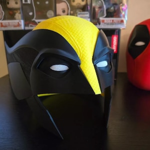 May include: A black and yellow plastic mask with a mesh eye covering. The mask is shaped like a superhero's head and has a yellow stripe down the middle. A red plastic mask with white eyes is in the background.