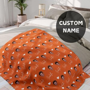 Puede incluir: Una manta naranja con un patrón repetido de caras, corazones blancos y el nombre "Isabella" en cursiva. La manta está colocada sobre una cama, mostrando un diseño personalizado.