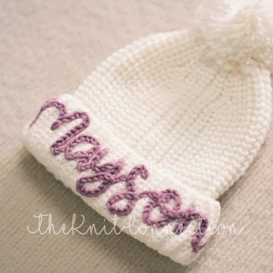 May include: White crocheted beanie with a pom pom on top. The beanie has the name "Mason" embroidered in purple yarn.