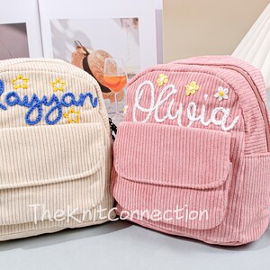 May include: Two small corduroy backpacks with embroidered names. The backpack on the left is beige with the name "Raysyan" embroidered in blue. The backpack on the right is pink with the name "Olivia" embroidered in white.