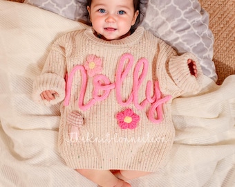May include: A cream coloured knitted jumper with the name "Molly" spelled out in pink yarn. The jumper has pink flower embellishments.