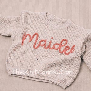 May include: A white knitted sweater with a pink embroidered name "Maidel" on the front. The sweater has a speckled texture and a crew neck.